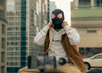 Person in superhero mask sitting on urban rooftop with camera equipment.