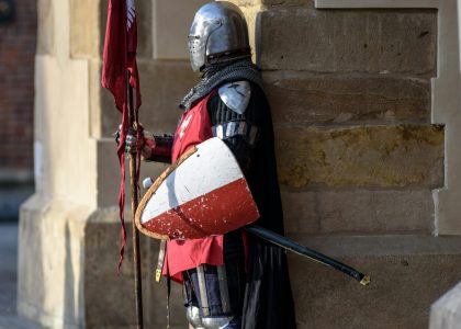 A medieval knight in armor stands guard with a shield and flag outside a stone building.